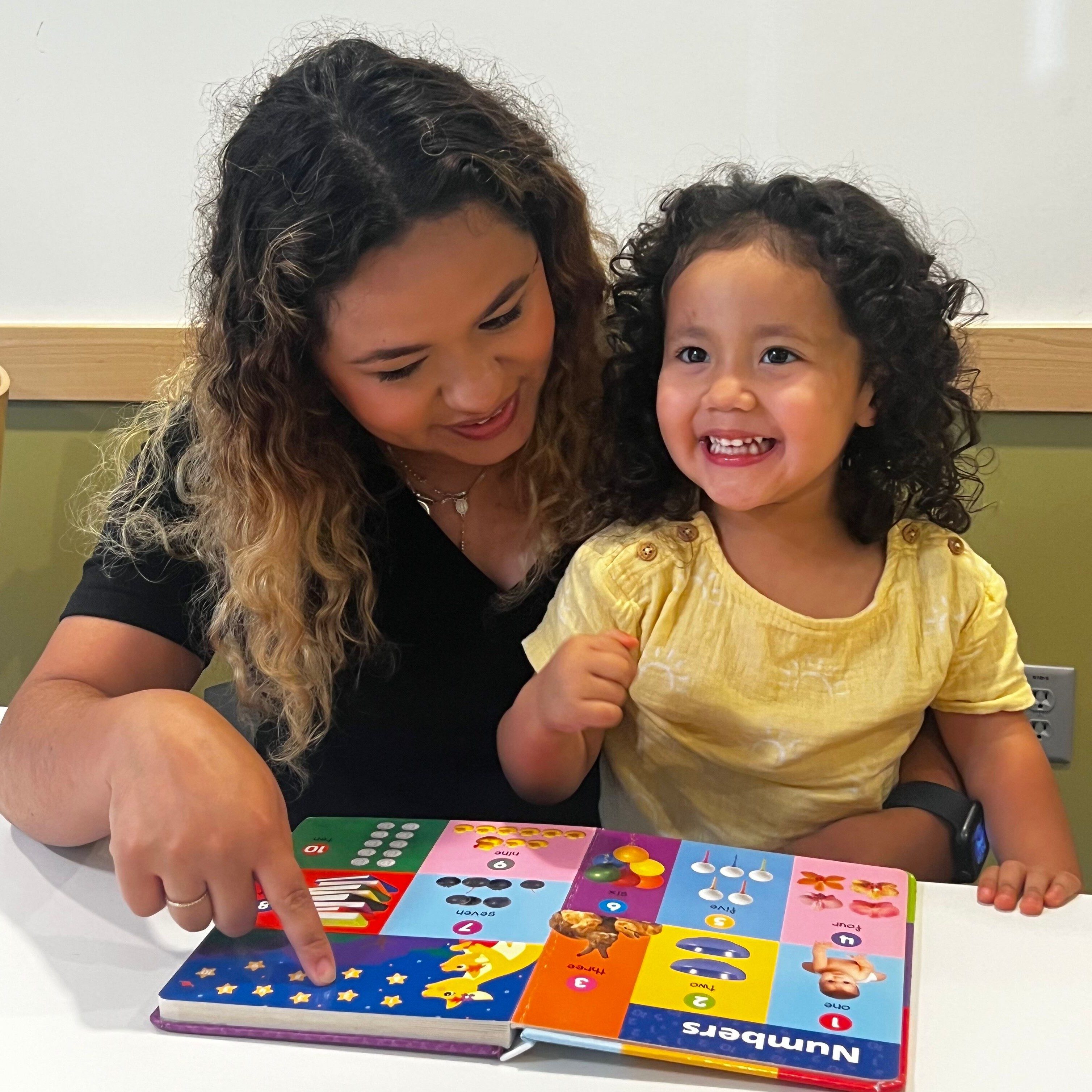 A smiling woman looking down and pointing to a book sitting next to a little girl smiling