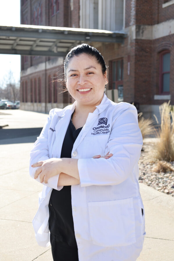 Dental Hygienist Mayra Perez wears a white coat and smiles while standing in front of a OneWorld building.