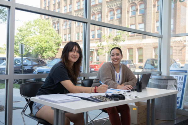 Members of the Patient Support Team smile from behind a check-in table. A window in the background shows OneWorld buildings and a parking lot.