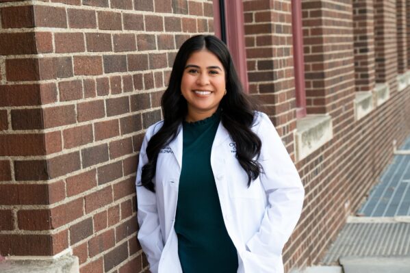 OneWorld Pediatric Nurse Practitioner Jiosajandy Garcia-Reyna, MSN, APRN, CPNP-PC, wears a white coat and smiles, while leaning against a brick wall.
