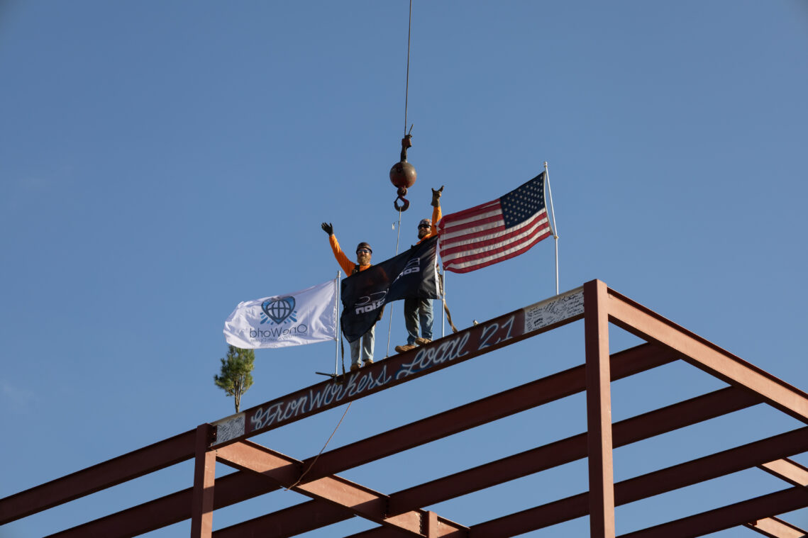 Two construction workers stand high along the top of a beam, which is the final beam of a future OneWorld building. OneWorld and Ronco Construction flags fly alongside the American flag.