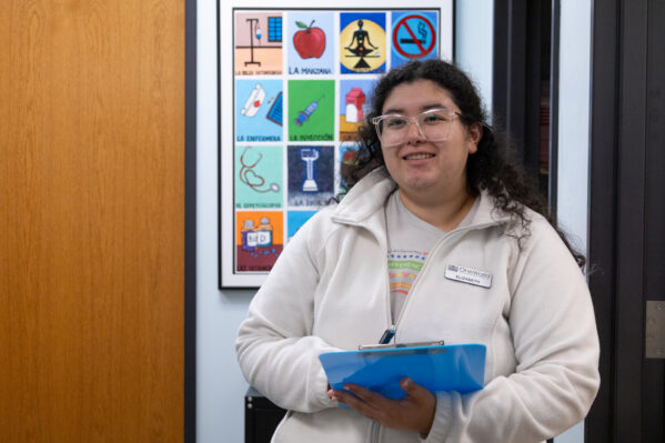 Medical Assistant Elizabeth Chavez-Troester smiles while holding a clipboard. Behind her is a colorful poster of health images. 