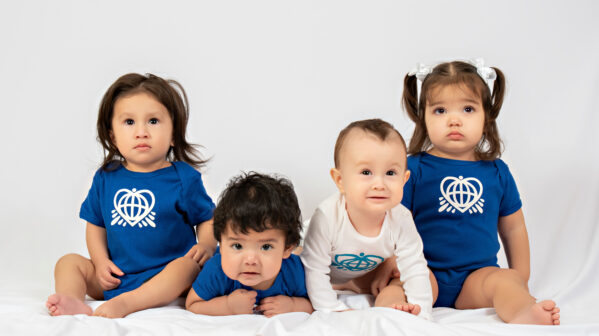 Four very cute babies sit in a line. They are wearing OneWorld onesies.
