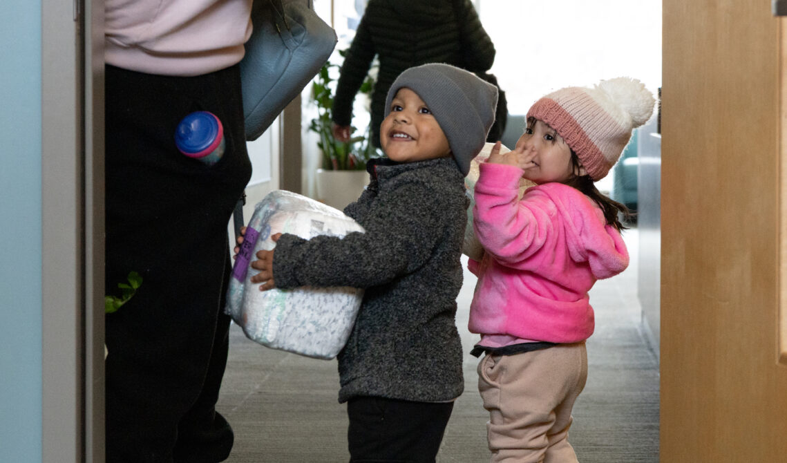 Two toddler smile while walking through a doorway. One child holds a package of diapers.
