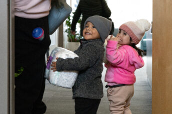Two toddler smile while walking through a doorway. One child holds a package of diapers.